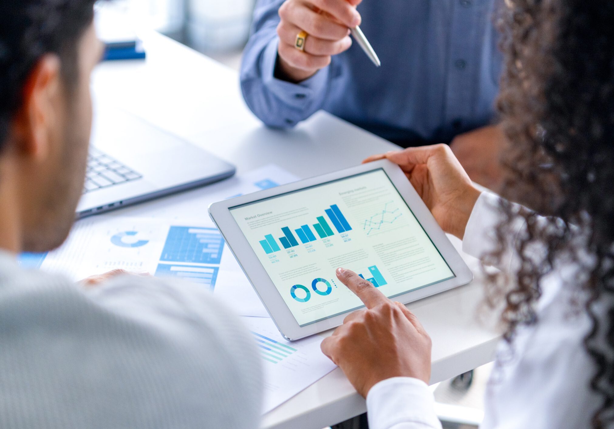 Close up of three people looking at financial data with graphs and charts.  All their hands can be seen and one person is pointing with a pen. There is paperwork on the desk showing more finance information